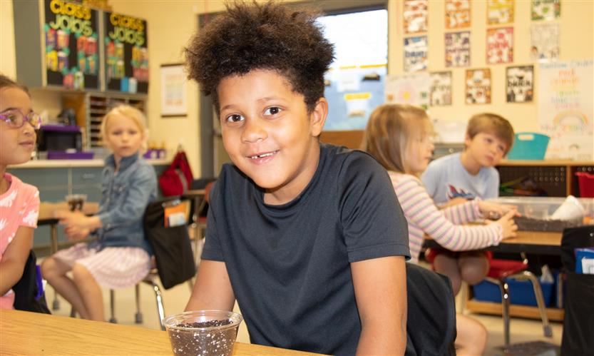 A boy smiles with a small plastic cup filled with dirt on the table in front of him.